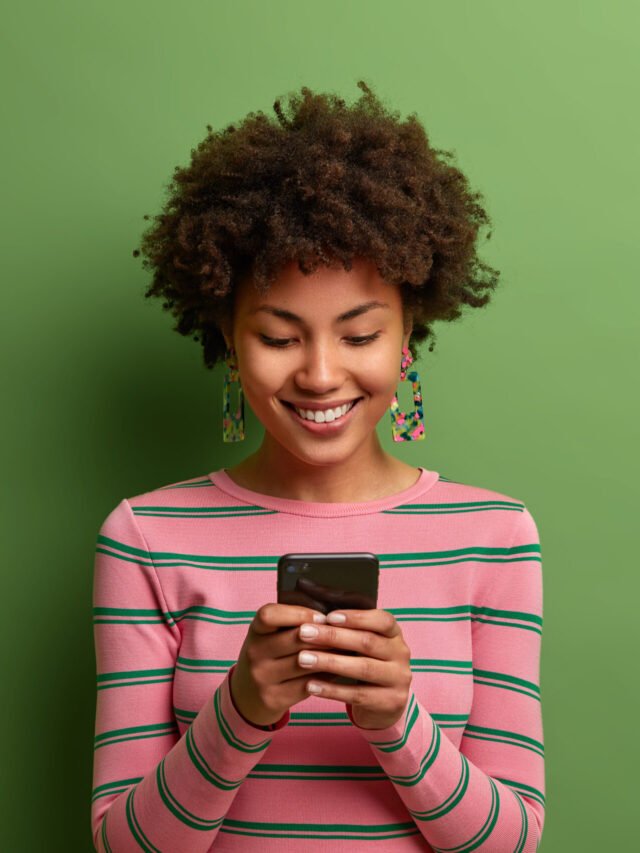 Pretty curly haired woman in striped jumper uses new application on smartphone enjoys high speed internet and types messages. Glad dark skinned teenager uses modern device isolated on green wall