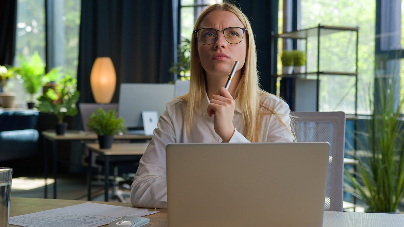 Mulher jovem pensativa usando óculos em frente ao laptop em ambiente de escritório moderno, representando profissional da Geração Z no trabalho
